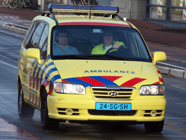 Ein gelber Krankenwagen mit zwei sichtbaren Insassen fährt an parkenden Fahrr√°dern auf einem Gehweg vorbei, mit einem Glasfenster-Geb√°ude im Hintergrund.