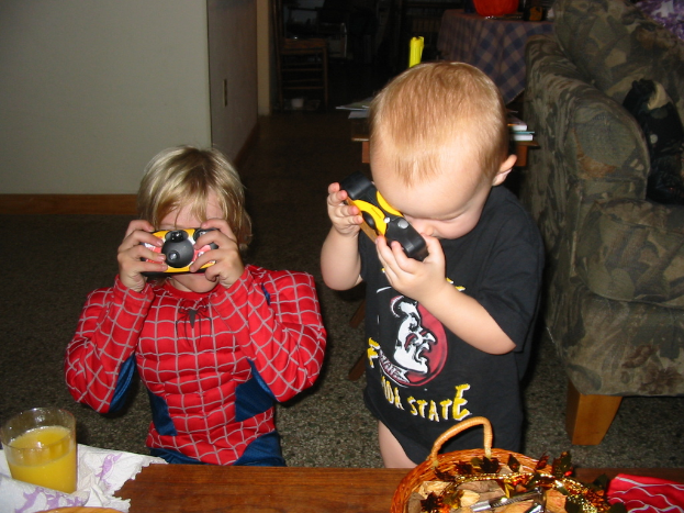 Zwei Kinder, eines im Spider-Man-Kostüm, halten eine Spielzeugkamera; ein hölzerner Tisch mit einem Glas und einem Korb ist im Vordergrund, eine Couch ist rechts und ein weiterer hölzerner Tisch ist im Hintergrund.