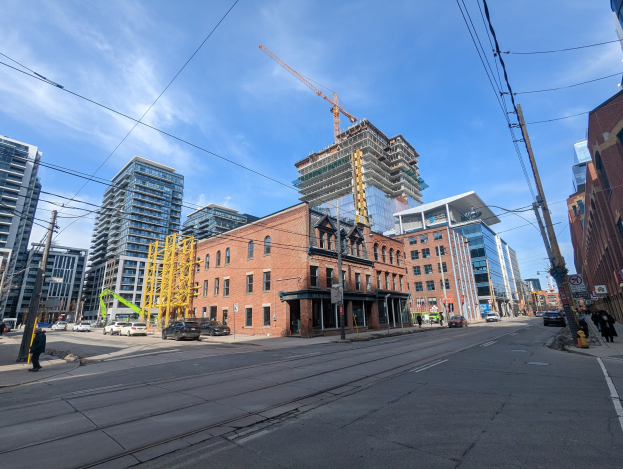 Eine belebte Straße in Toronto mit Fahrzeugen, Füpßlern, Oberleitungen, Schildern und Gebäuden, darunter eine Baustelle mit einem Kran im Hintergrund unter einem bewölktem Himmel.
