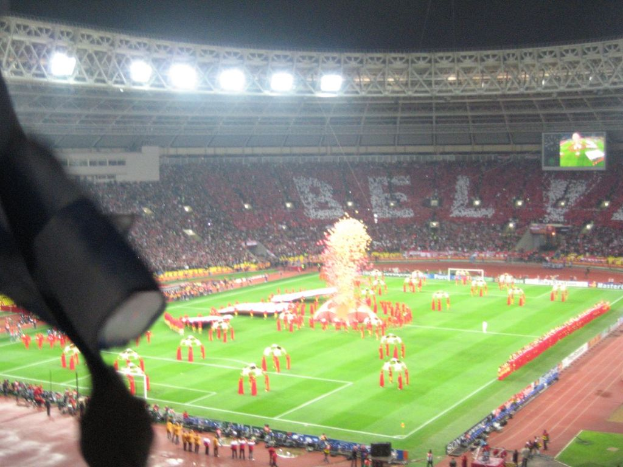 Ein Stadion mit Flutlicht, Eisengittern, Zuschauern, stehenden Personen, Werbetafeln, Bildschirmen, Überwachungskamera und Fahnen und Dekorationen.