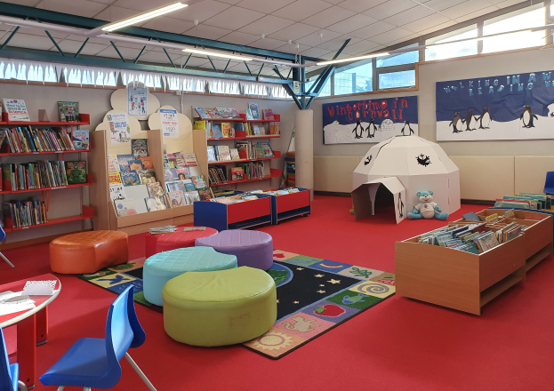 Eine hell erleuchtete Kinderbibliothek mit Bücherregalen, Tischen, Stühlen, Teppichen, Wanddisplays und Deckenleuchten.