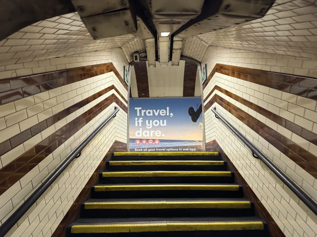 Eine Treppe in einer U-Bahn-Station mit einem Schild "Reise, wenn du wagst" oben, flankiert von Geländern und StationWsänden im Hintergrund.