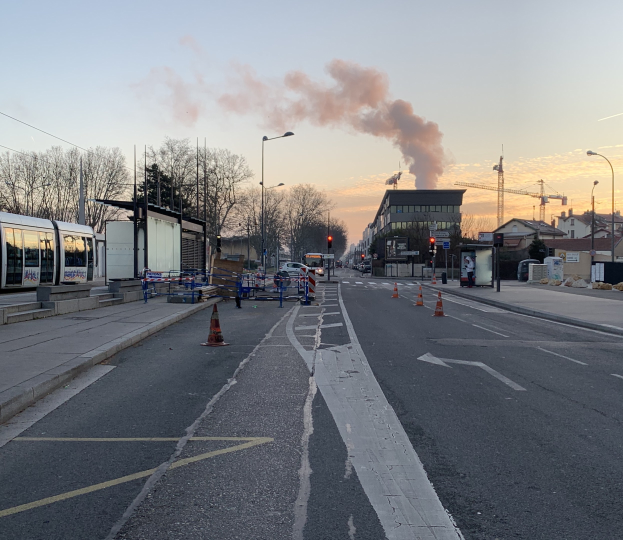 Straßenszene mit Verkehrskegeln, einem Bus, Laternenmasten, Bäumen, Gebäuden, Kränen und Rauch, der aus einem entfernten Gebäude aufsteigt.