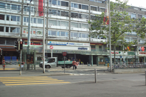 Eine Straßenansicht mit einem Lastwagen, der an hohen Gebäuden vorbeifährt, Fußgängern auf dem Bürgersteig, parkenden Fahrrädern, Verkehrsampeln, Schildern, Strommasten, Drähten, einem Baum und einem bewölkten Himmel.