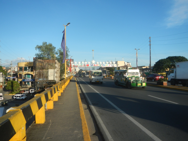 Eine belebte Stadtstraße mit Fahrzeugen, einer Mitteltrennung, einem Fahnenmast mit Flagge, Gebäuden, Bäumen, Strommasten mit Drähten und einem bewölkten Himmel.