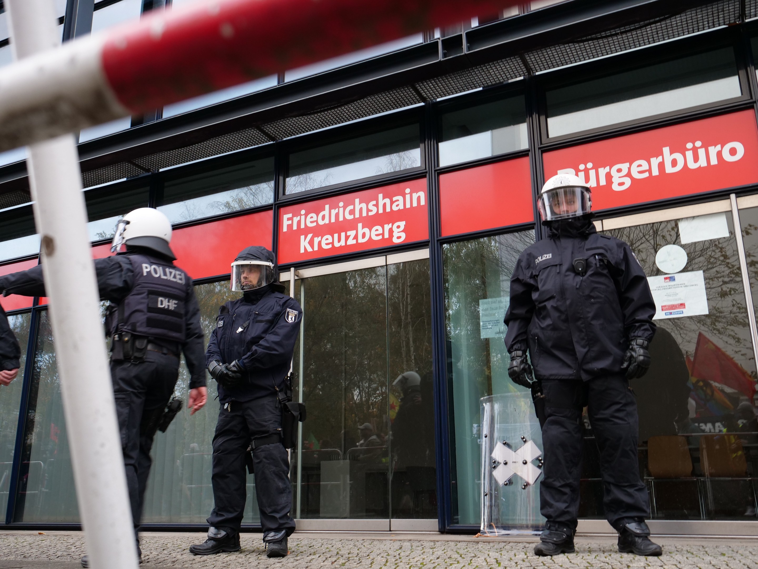Polizisten in Uniform vor einer Glaswand stehend, links ein Mast und Schilder mit 'Friedrichshain Kreuzberg' Text im Hintergrund.
