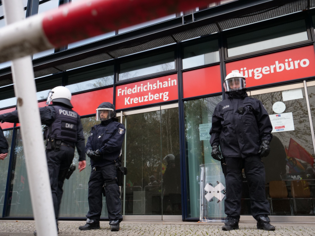 Polizisten in Uniform vor einer Glaswand stehend, links ein Mast und Schilder mit 'Friedrichshain Kreuzberg' Text im Hintergrund.