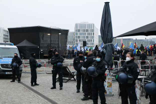 Polizeibeamte in Einsatzausrüstung stehen hinter Barrieren und blicken auf eine Menge von Demonstranten, die Fahnen schwenken und Helme und Masken tragen.