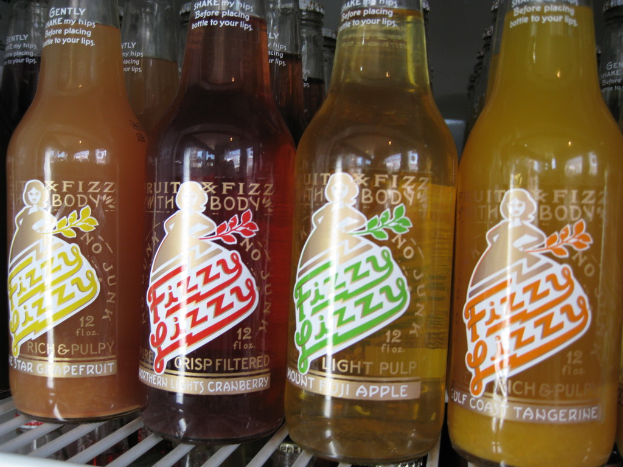 Drei farbenfrohe Getränkeflaschen mit der Aufschrift 'Fizzy Lizzy' in Gelb, Rot und Orange auf einem Tisch.