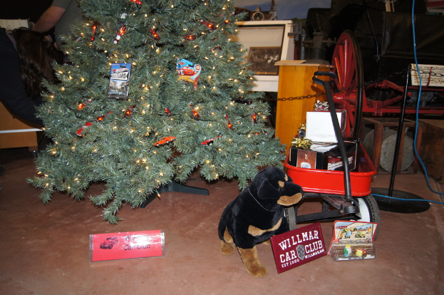 Eine Szene mit einem Hundespielzeug, Namensschildern, einem Wagen mit Gegenständen, einem Weihnachtsbaum, einem Rad, einem Pfahl, einer Kette, einer Holzplatte, einer Person, die steht, und verschiedenen anderen Gegenständen im Hintergrund.