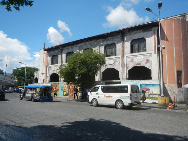 Eine belebte Stadtstraße mit Fahrzeugen, Fußgängern, Bäumen, Laternenpfählen, einem großen Gebäude mit Wandgemälde, einer Brücke im Hintergrund und einem bewölkten Himmel.