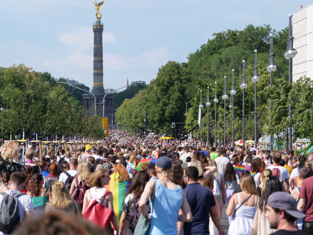 Eine große Menschenmenge geht eine Straße in Berlin, Deutschland, entlang, mit Laternenpfählen am Weg, Bäumen und einem Turm mit Statue im Hintergrund und umliegenden Gebäuden unter einem bewölkten Himmel.