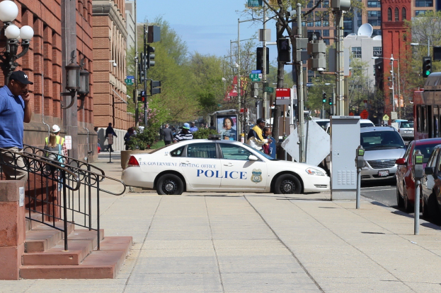 Eine belebte Stadtstraße mit einem parkenden Polizeiwagen, fahrenden Fahrzeugen einschließlich eines Busses und eines Autos, Fußgängern auf dem Gehweg, Gebäuden mit Fenstern und Geländern, Laternenpfählen, Bäumen im Hintergrund und einem klaren blauen Himmel.