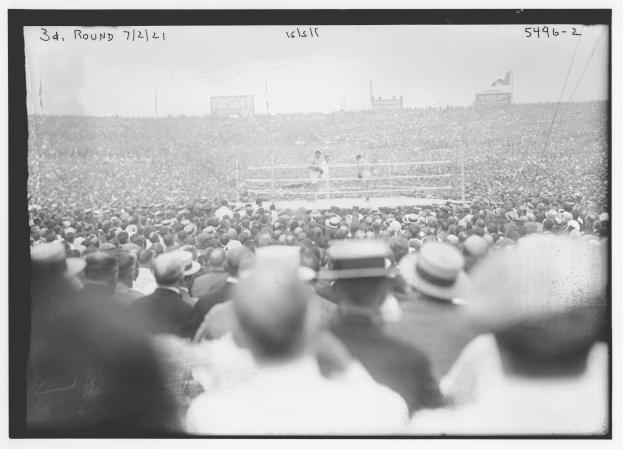 Ein Schwarz-Weiß-Foto einer Menge, die einen Boxkampf in einem Stadion verfolgt, mit zwei Personen im Ring, umgeben von Menschen mit Hüten, und Zahlen oben und unten auf dem Foto.