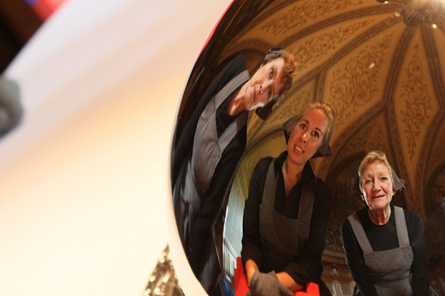Ein reflektierender Ball auf einem weißen Tisch zeigt das Spiegelbild von drei lachenden Frauen, die leicht gebückt sind, mit einem Dach im Hintergrund.