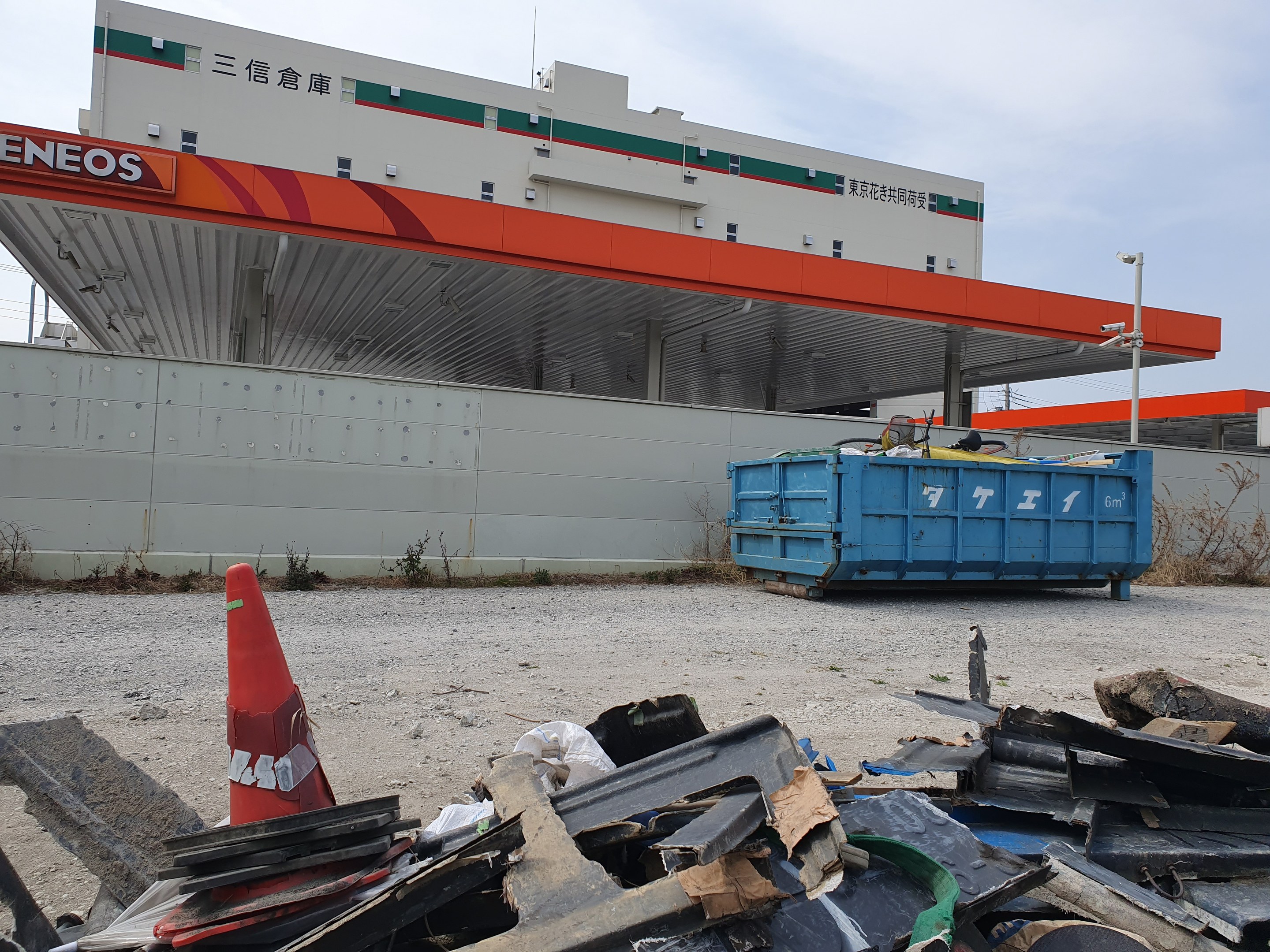 Großes Gebäude mit Stapeln von Schrott, einem Verkehrskegel und einem blauen Container davor, umgeben von Pflanzen, Pfählen und Himmel während der Abrissarbeiten.