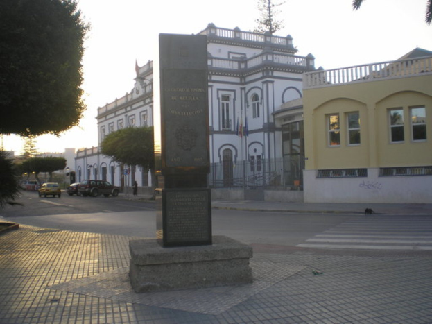 Ein Denkmal steht in der Mitte mit HÃ¤usern, Fahrzeugen auf einer StraÃŸe, BÃ¤umen links, Himmel Ã¼ber und Gehweg unten.