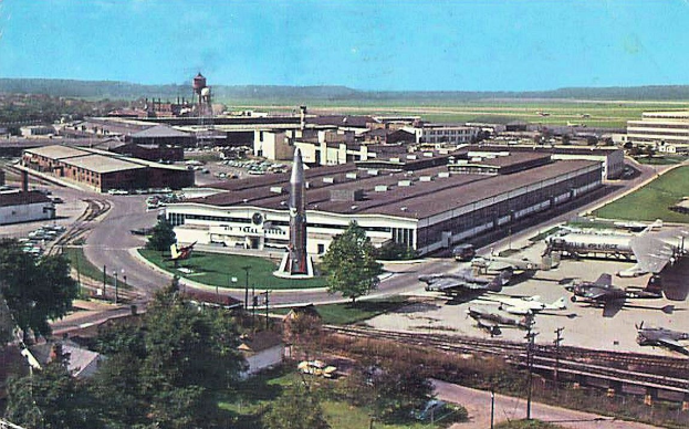 Alte Schwarz-Weiß-Airport-Szene mit geparkten Flugzeugen, Gebäuden, Bäumen, Straßen, Fahrzeugen, Versorgungsmasten, Gras, Bahnschienen, fernen Türmen, Hügeln und einem klaren blauen Himmel, mit Text entlang der unteren Kante.