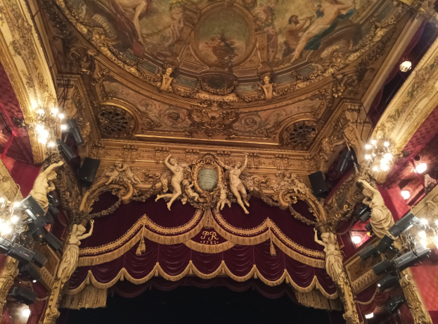 Innenansicht eines Theaters mit rotem Vorhang, Kronleuchtern, Skulpturen, Lampen und einer Deckenmalerei, mit einer Bühne und ein paar Menschen davor.
