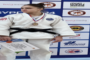 Eine Frau in einem weißen Judo-Anzug mit einem schwarzen Gürtel steht auf einem Podium, hält eine Medaille und ein Zertifikat und hat eine logo-bedeckte Tafel im Hintergrund.