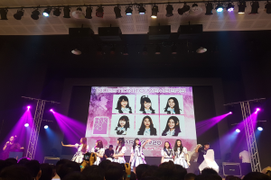 Eine Gruppe von Menschen performs auf einer hell erleuchteten Bühne mit Trägern, während ein Sänger ein Mikrofon auf der linken Seite hält und ein anderer ein Instrument spielt, während eine Menge mit Kameras unten und ein großer Bildschirm im Hintergrund Bilder der K-Pop-Gruppe BK48 anzeigt.