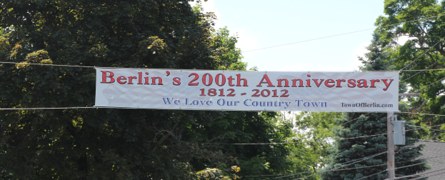 Ein Banner mit der Aufschrift 'Berliner Jubiläum 200' hängt an einem Draht in der Vordergrund, mit Bäumen und einem Haus im Hintergrund unter einem klaren Himmel.