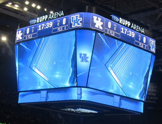 Großer Bildschirm auf einem Basketballfeld mit der Aufschrift "Kentucky Wildcats vs Florida State Seminoles" und Zuschauern auf den Tribünen und Beleuchtung von oben.