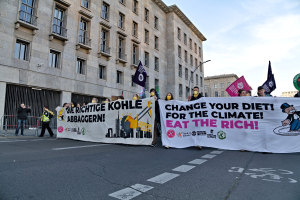 Gruppe von Menschen mit einem Banner 'Verändere deine Ernährung für das Klima, iss die Reichen' auf einer städtischen Straße mit Gebäuden und Laternen unter einem bewölkten Himmel.