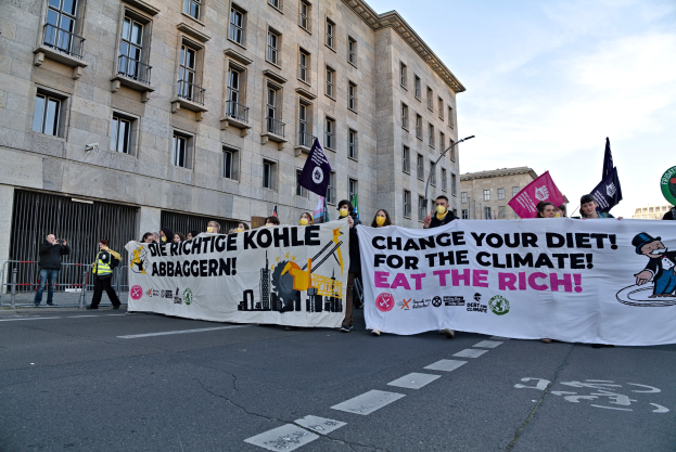 Gruppe von Menschen mit einem Banner 'Verändere deine Ernährung für das Klima, iss die Reichen' auf einer städtischen Straße mit Gebäuden und Laternen unter einem bewölkten Himmel.