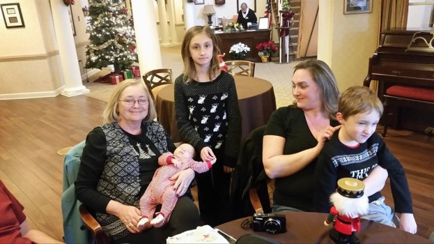 Ein Raum mit einer Wand, die ein Fotoframe, einen Weihnachtsbaum, eine Lampe und Blumenpflanzen zeigt, eine Treppe, die nach oben führt, sowie einige Menschen an Tischen mit einem Spielzeug und einer Kamera auf einem Tisch.