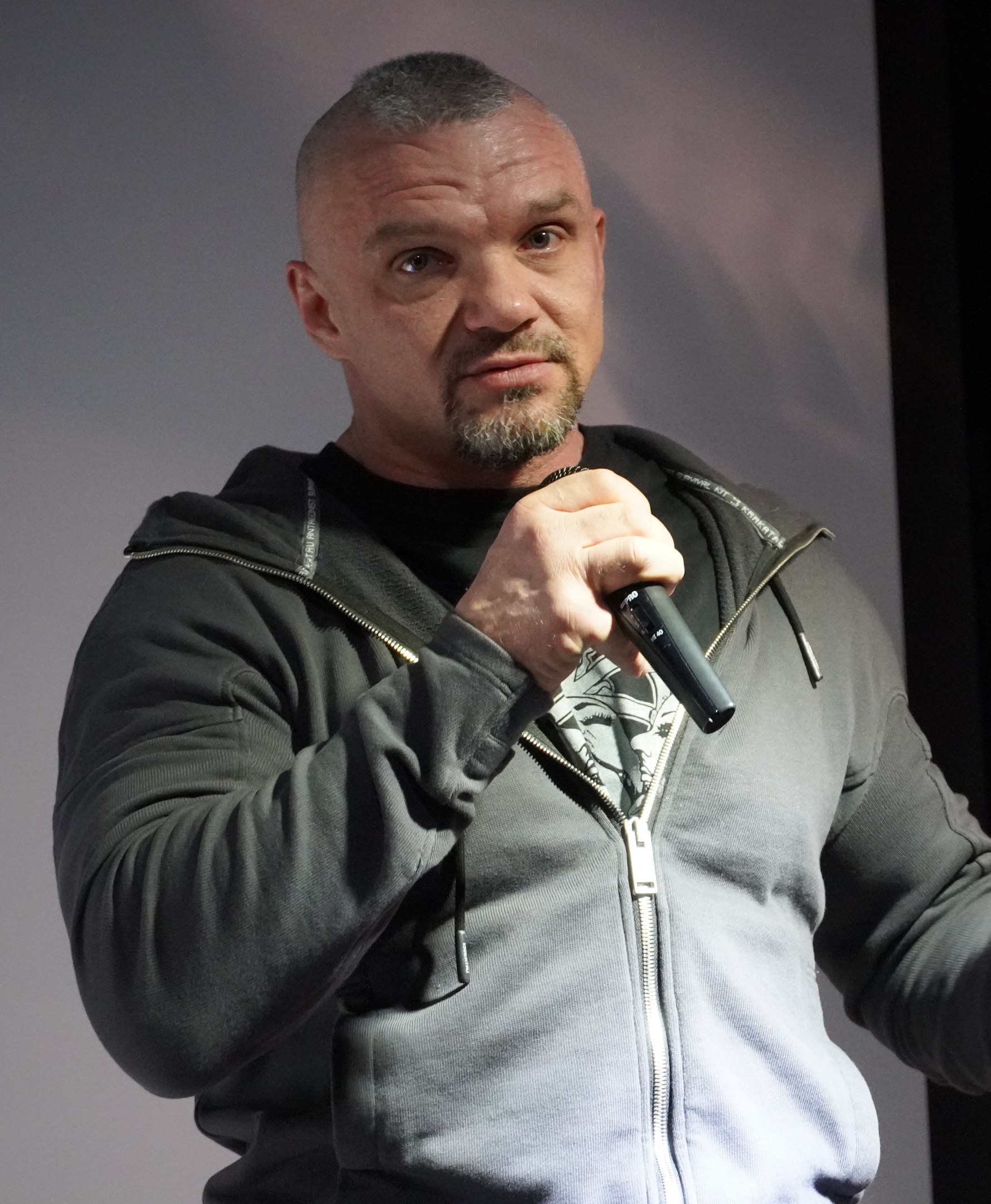 Ein Mann in einer schwarzen Kapuzenjacke und einem schwarzen Anzug mit einem weißen Hemd hält ein Mikrofon und steht selbstbewusst vor einer Wand, wobei er wie ein Komiker wirkt.