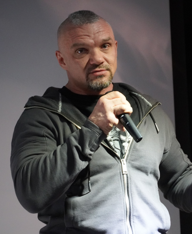 Ein Mann in einer schwarzen Kapuzenjacke und einem schwarzen Anzug mit einem weißen Hemd hält ein Mikrofon und steht selbstbewusst vor einer Wand, wobei er wie ein Komiker wirkt.