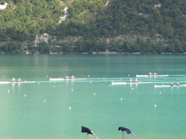 Gruppe von Menschen bei einem Ruderwettbewerb auf einem Gewässer mit Bäumen und einem Hügel im Hintergrund, Kleider über Stühle drapiert.