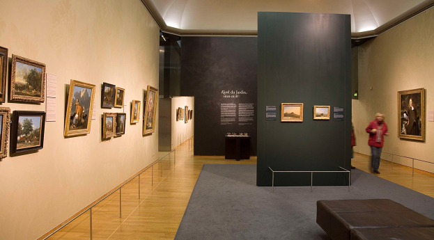 Ein großer Raum voller Gemälde an den Wänden, mit zwei Personen auf dem Boden, einer Couch, einem Tisch mit verschiedenen Gegenständen, einer Tafel mit dem Text "Karel du Jardin Ausstellung in der Nationalgalerie" und einer Decke mit Lampen.