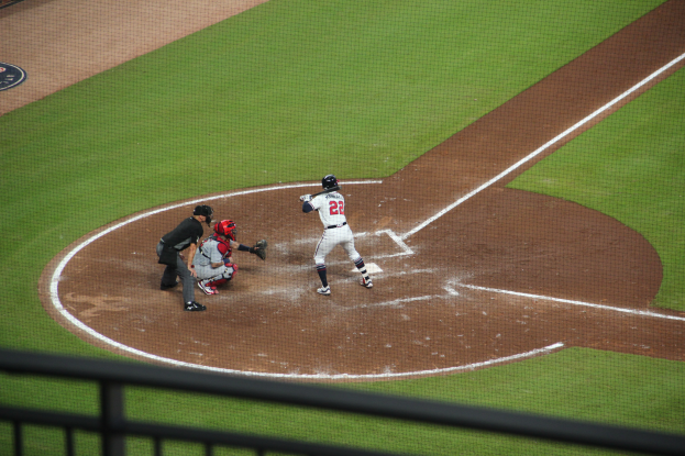 Ein Baseballspieler in einem Sportoutfit schwingt einen Schläger auf einem grünen Feld, wobei ein Geländer am unteren Bildrand zu sehen ist.