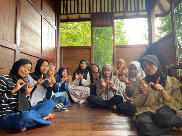 Eine Gruppe von Frauen sitzt auf dem Boden vor einer hölzernen Wand mit verstreuten Taschen und Gegenständen, die an einer Trainingssitzung in Indonesien teilnehmen, sichtbar durch Glastüren, die Bäume und Himmel zeigen.