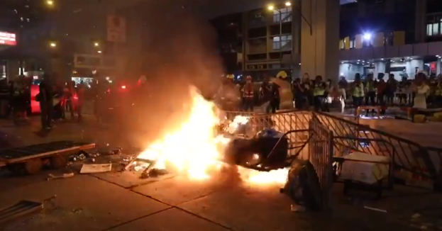 Eine Gruppe von Menschen steht um ein Feuer in der Mitte einer Straße, umgeben von Barrikaden und anderen Gegenständen, mit Gebäuden, Laternenmasten und einer Tafel im Hintergrund unter dem Nachthimmel.