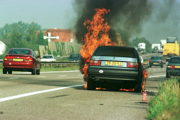 Ein Auto, das in Flammen steht, am Straßenrand mit anderen Fahrzeugen in der Nähe, Bäumen, Gebäuden und einem klaren blauen Himmel im Hintergrund und Gras mit einem Feuerlöscher auf der rechten Seite.