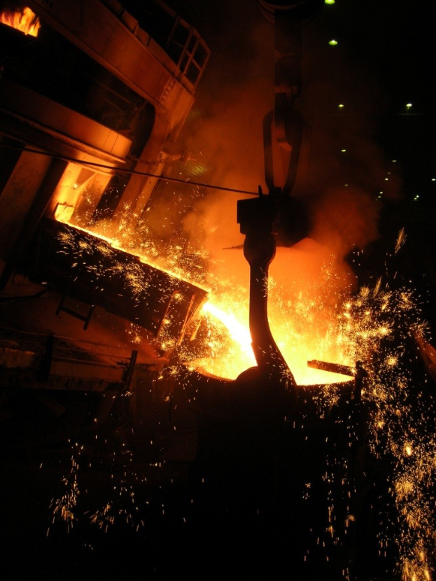 Ein Stahlwerk bei Nacht mit einem Kran, der ein großes Stück Stahl in einen Ofen hebt, dessen Feuer den Bereich mit einem hellen orangen Glühen erhellt, das sich vor einem dunklen Hintergrund abhebt.