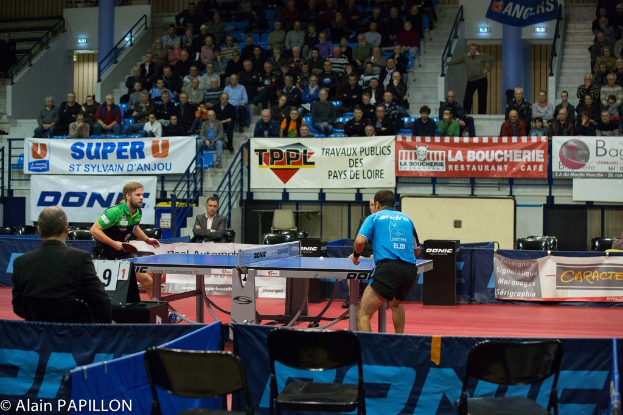 Zwei Männer spielen drinnen Tischtennis, mit mehreren Zuschauern, die stehen und sitzen, einigen leeren Stühlen und sichtbaren Werbetafeln, mit Text in der unteren linken Ecke.