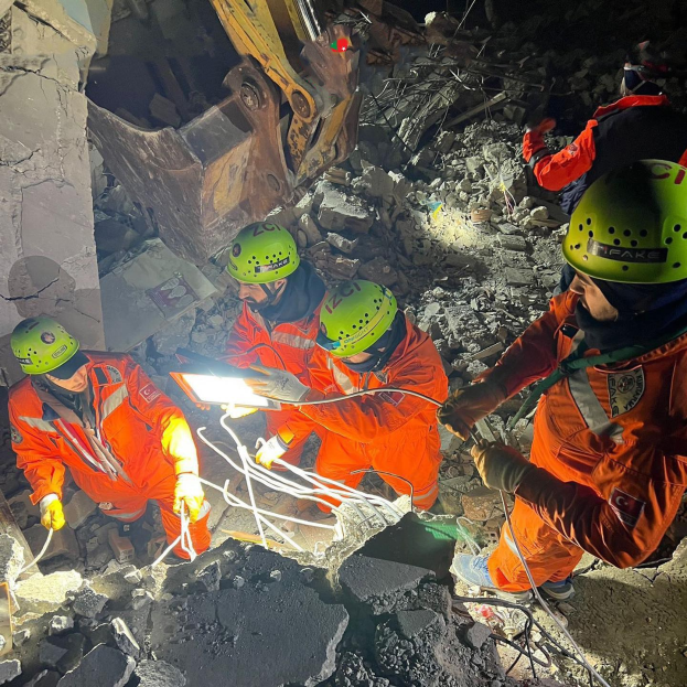 Eine Gruppe von Rettungskräften in orangefarbenen Uniformen und grünen Helmen beseitigt Trümmern von einem eingestürzten Gebäude mit einem Kran im Hintergrund.