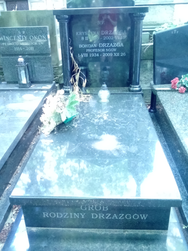 Ein Friedhof mit zwei benachbarten Grabsteinen, umgeben von Blumensträußen und anderen Gegenständen, mit einem Gebäude im Hintergrund.