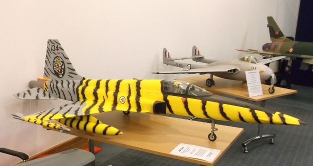 Verschiedene Flugzeugmodelle sind auf Tischen mit Etiketten angeordnet, ein Stuhl ist auf der linken Seite und eine Wand im Hintergrund zu sehen.