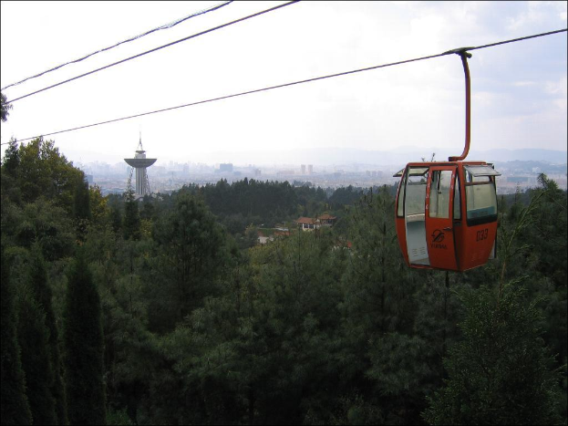 Eine Seilbahn bewegt sich im Vordergrund mit einer Stadtlandschaft, einschließlich Gebäuden, einem Turm und Bäumen, unter einem bewölkten Himmel im Hintergrund.