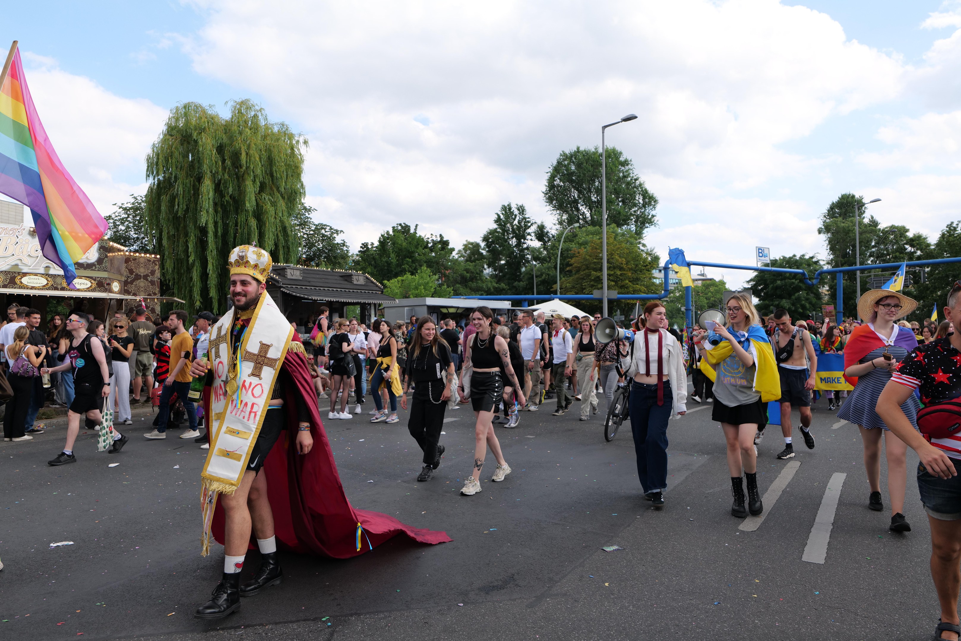 Eine Gruppe von Menschen marschiert bei der Gay Pride Parade 2018 mit einer Regenbogenfahne und Musikinstrumenten, umgeben von Laternenmasten, Bäumen, Hütten und einem bewölkten Himmel.
