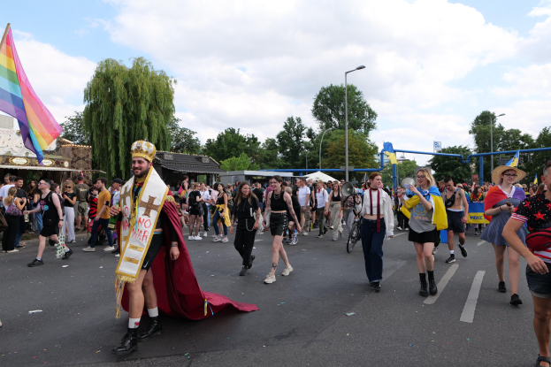 Eine Gruppe von Menschen marschiert bei der Gay Pride Parade 2018 mit einer Regenbogenfahne und Musikinstrumenten, umgeben von Laternenmasten, Bäumen, Hütten und einem bewölkten Himmel.
