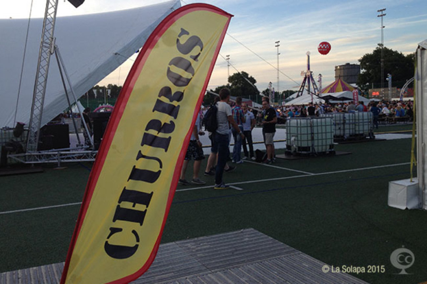 Eine KarnevalsSzene mit Zelten, einem zentralen Mast, Menschen, Bäumen, Strommasten, Straßenbahnen auf der Straße und einem Eisenständer auf der linken Seite, unter einem Himmel.