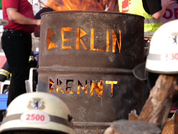 Menschen um ein Feuerhydrant mit der Aufschrift "Berlin" versammelt, brennender Eimer im Vordergrund, Helme unten und hölzernes Objekt rechts.