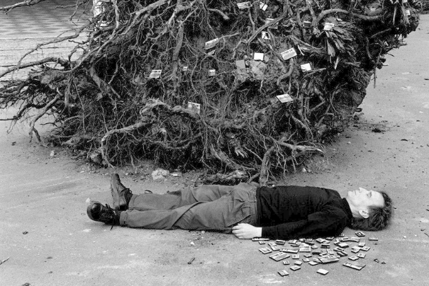 Ein Mann liegt neben einem Geldstapel auf dem Boden, trägt Wanderschuhe und hat einen Baum im Hintergrund, auf einem Schwarz-Weiß-Bild.