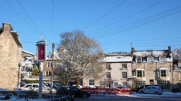 Fahrzeuge auf einer schneebedeckten Straße mit Häusern, Bäumen, Strommasten und schneebeladenen Drähten im Hintergrund.